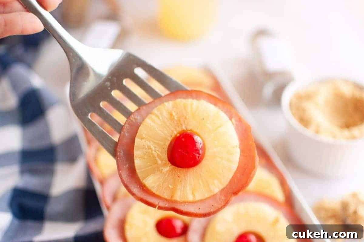 A metal spatula holds a slice of ham topped with a pineapple ring and a red maraschino cherry; more similar slices are visible in the background.