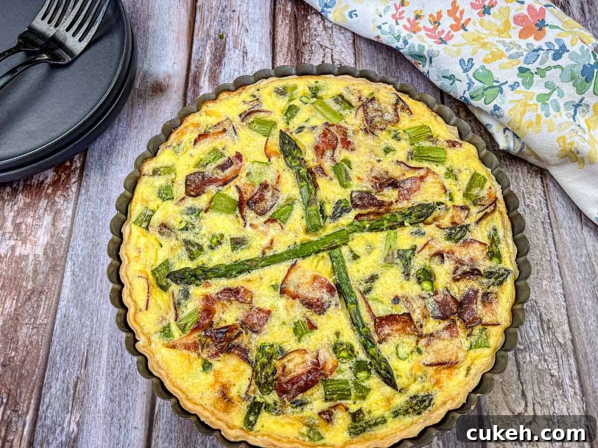 Asparagus & Ricotta Quiche in a round pan on a wooden table, with a floral napkin and black plates with forks nearby.
