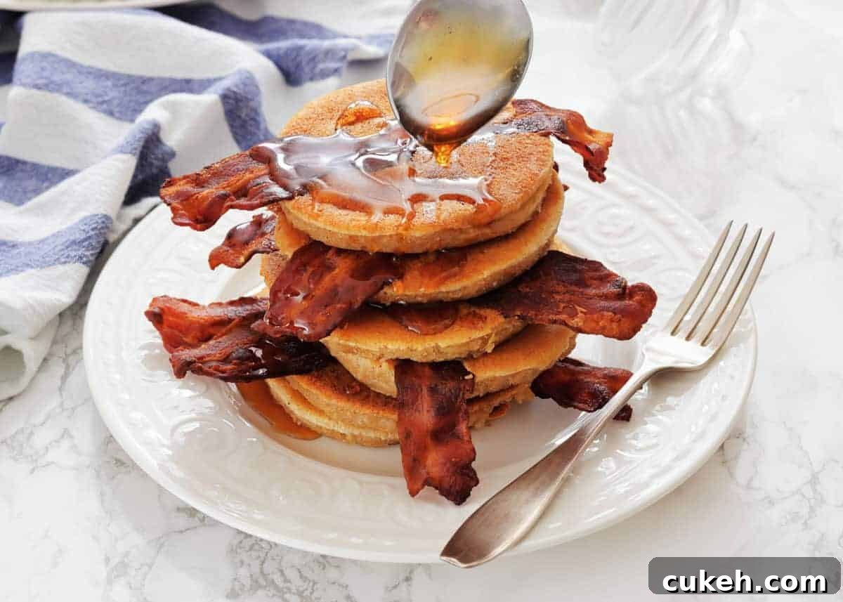 A stack of pancakes with strips of bacon, being drizzled with syrup from a spoon, on a white plate with a fork beside it.