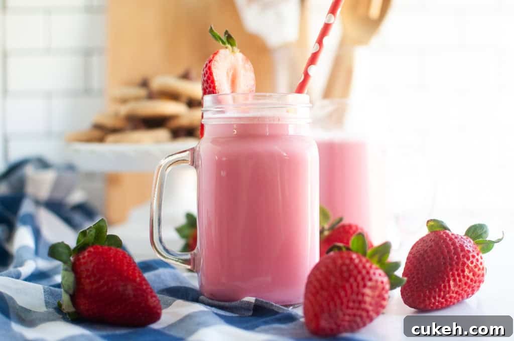 Two pink strawberry milk in glass jars with straws and strawberry garnishes, on a blue and white checkered cloth.