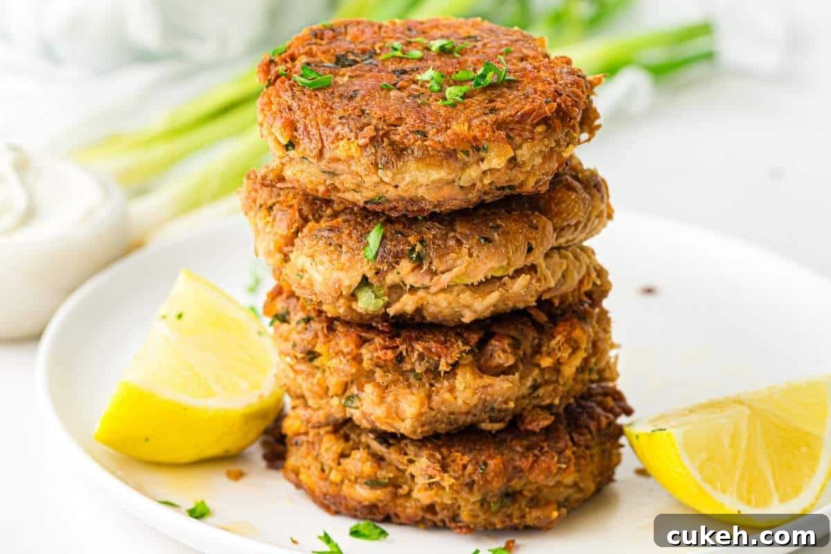 A stack of four golden-brown tuna patties garnished with chopped herbs, served on a white plate with two lemon wedges.