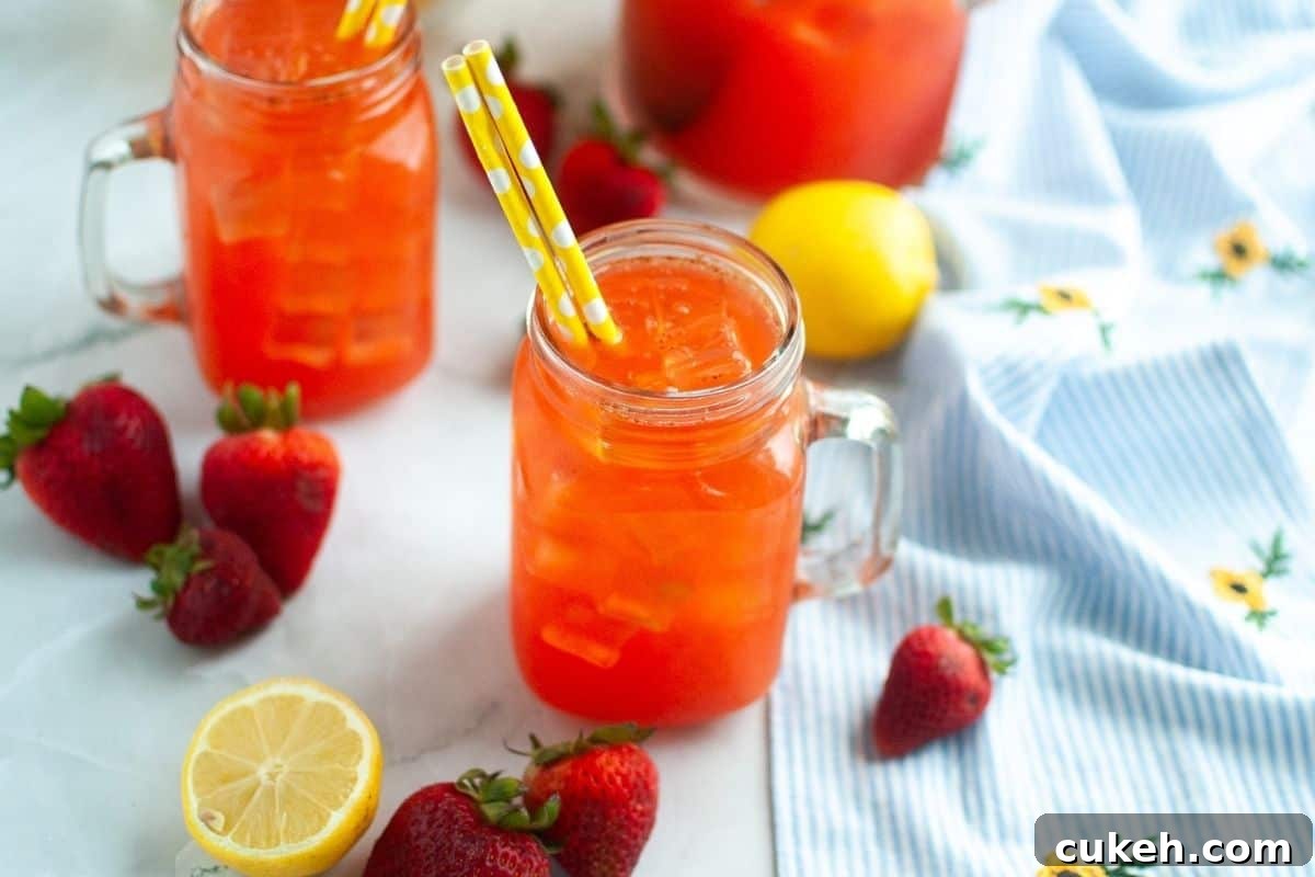 Glass mason jars filled with iced strawberry lemonade on a table, surrounded by fresh strawberries, a halved lemon, and a blue floral cloth.