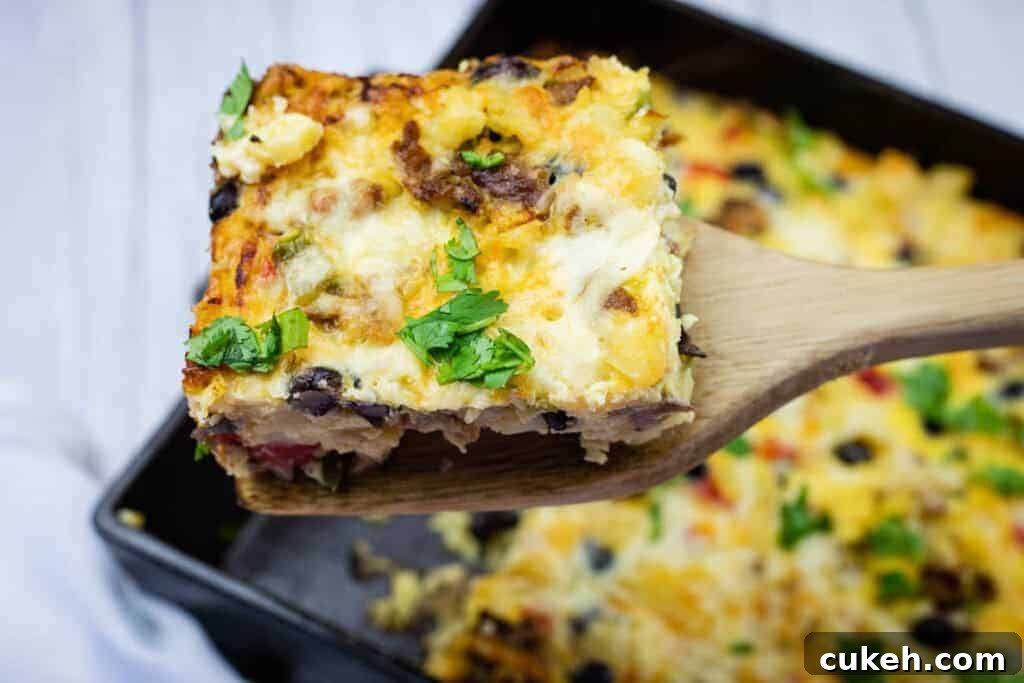 A wooden spatula holds up a square slice of baked casserole topped with melted cheese and chopped herbs, with more casserole in the background.