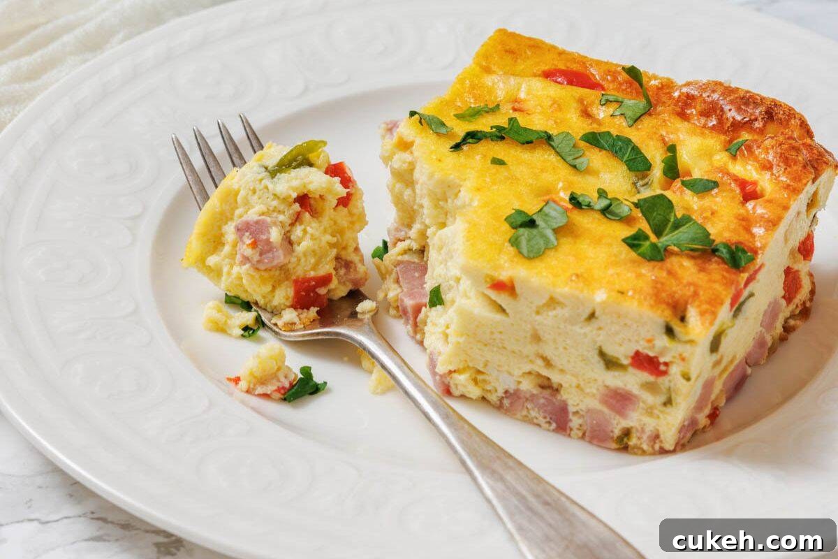 A slice of baked Western omelet with ham, red peppers, and herbs is served on a white plate with a fork holding a bite-sized piece.