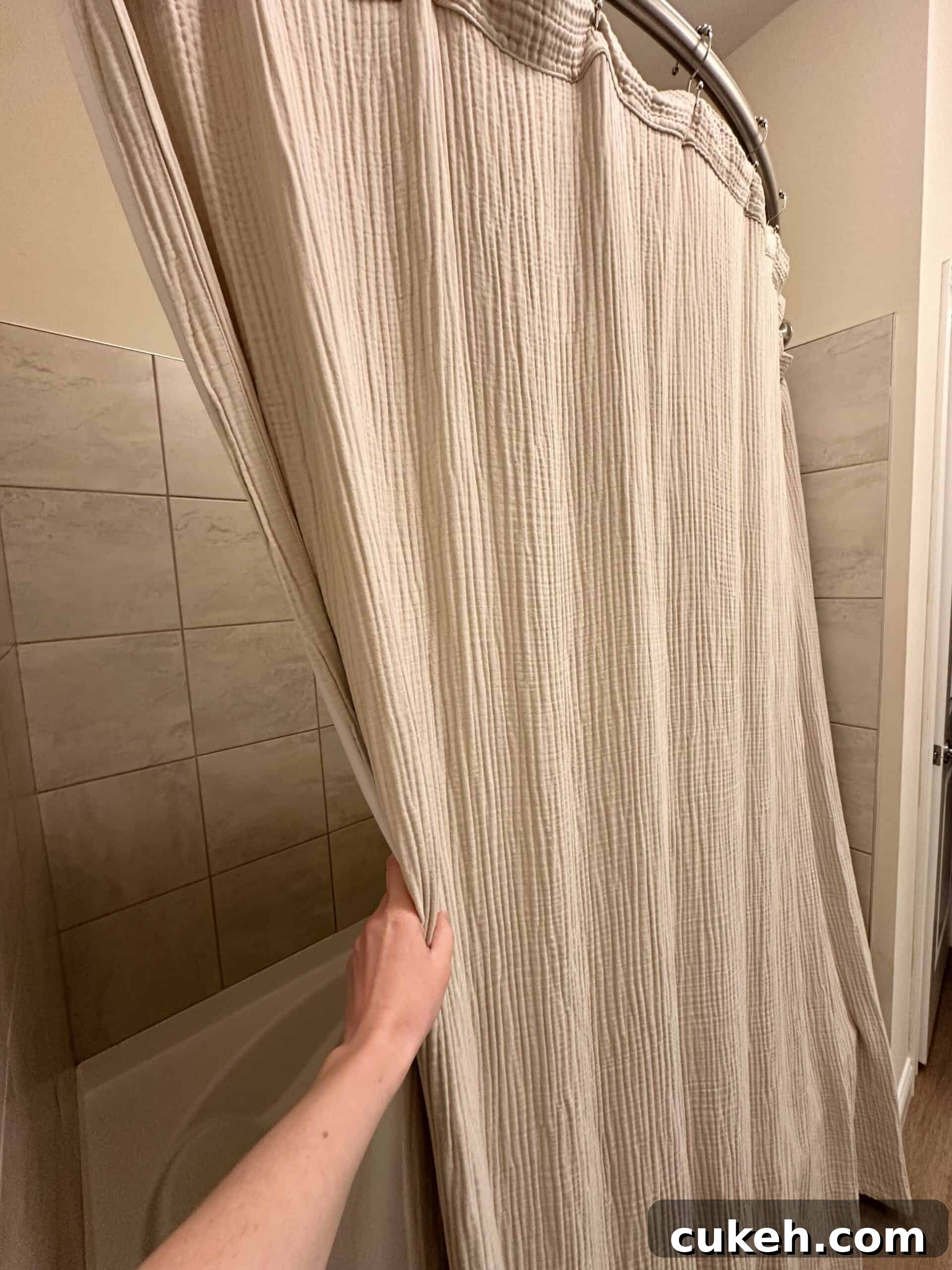 Hand pulling back a beige, textured cotton shower curtain in a tiled bathroom.