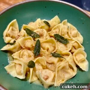 Butternut Squash Tortellini in Sage Brown Butter Sauce 4 A green bowl filled with tortellini, topped with crispy sage and a sprinkle of cheese, placed on a wooden surface. The pasta is golden brown, glazed with a light sauce.