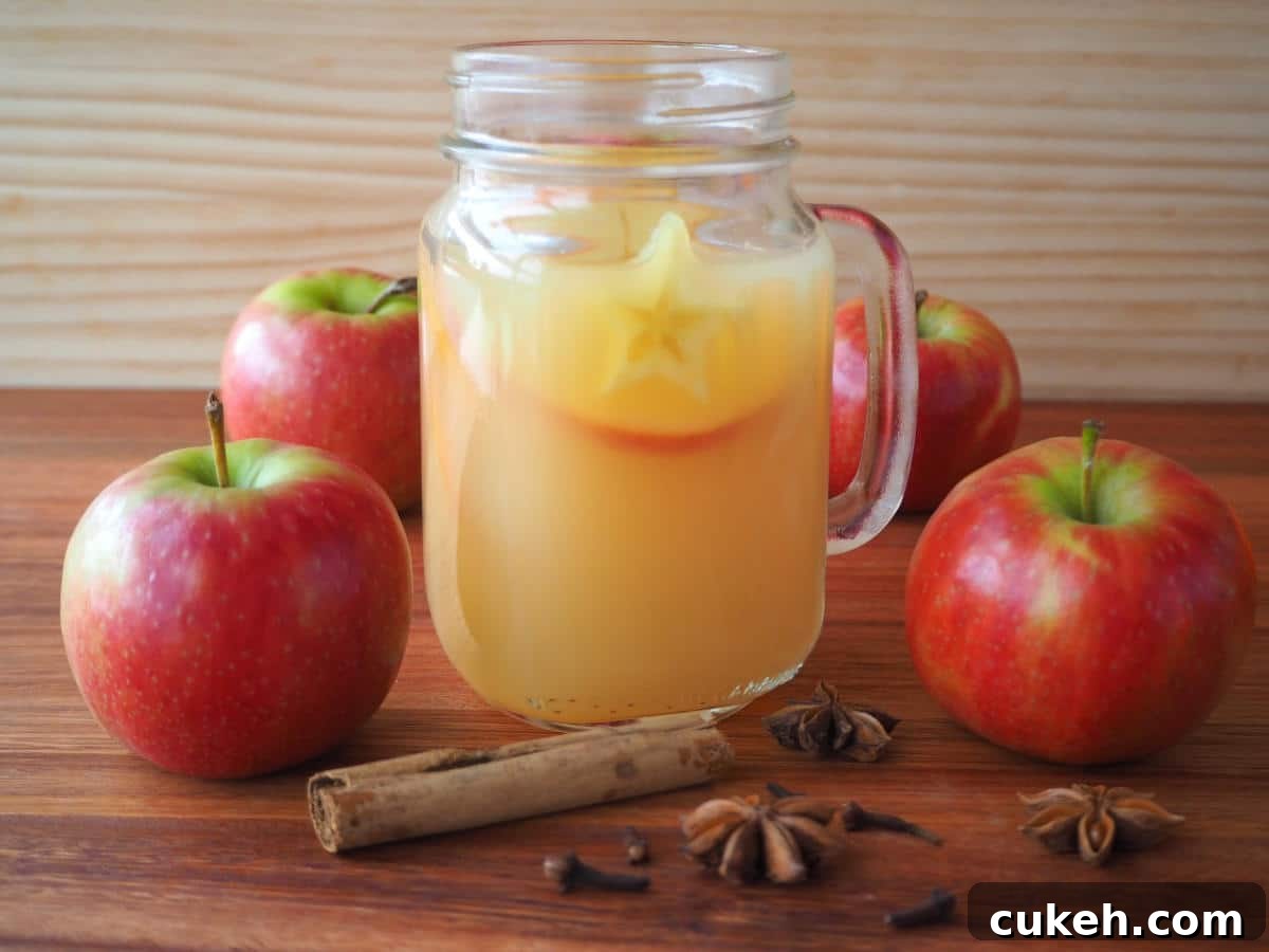 side view of a mason jar mug filled with mulled apple cider, with apples and spices on the side, on a brown board with a light brown background.