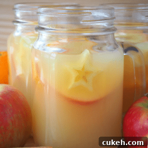 close up side view of a mason jar mug filled with mulled apple cider, with apples and spices on the side, on a brown board with a light brown background.