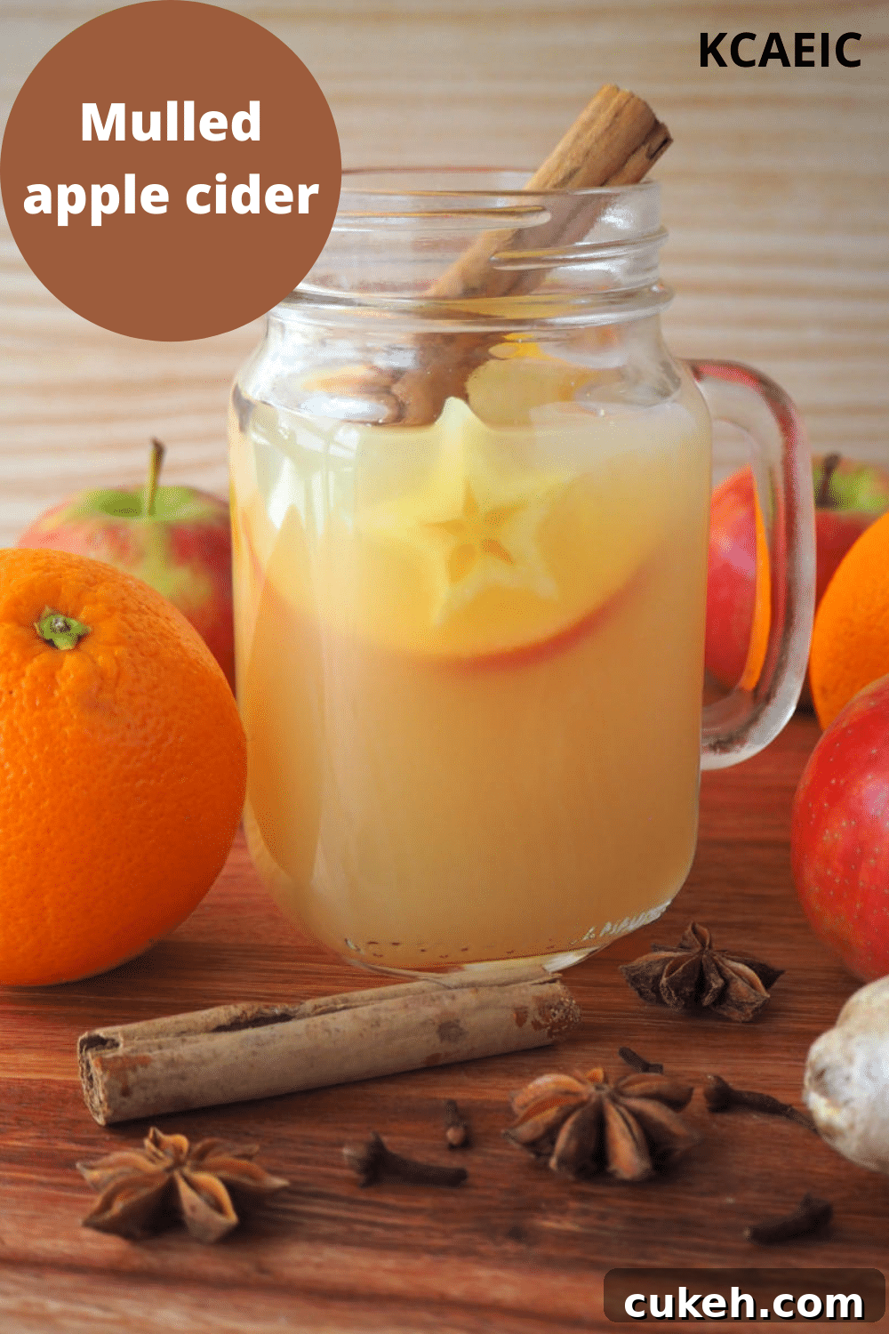 side view of a mason jar mug filled with mulled apple cider, with apples, oranges and spices on the side, on a brown board with a light brown background, with text overlay, mulled apple cider and KCAEIC.