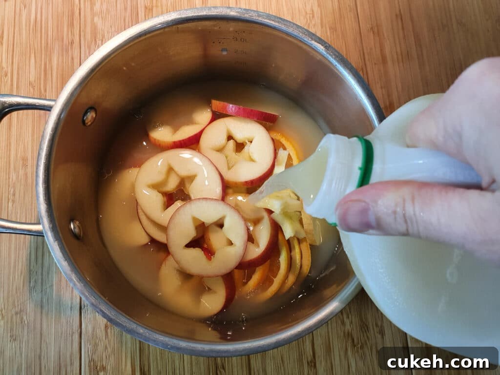 adding apple cider or cloudy apple juice to a pot containing sliced apples and oranges, on a chopping board.