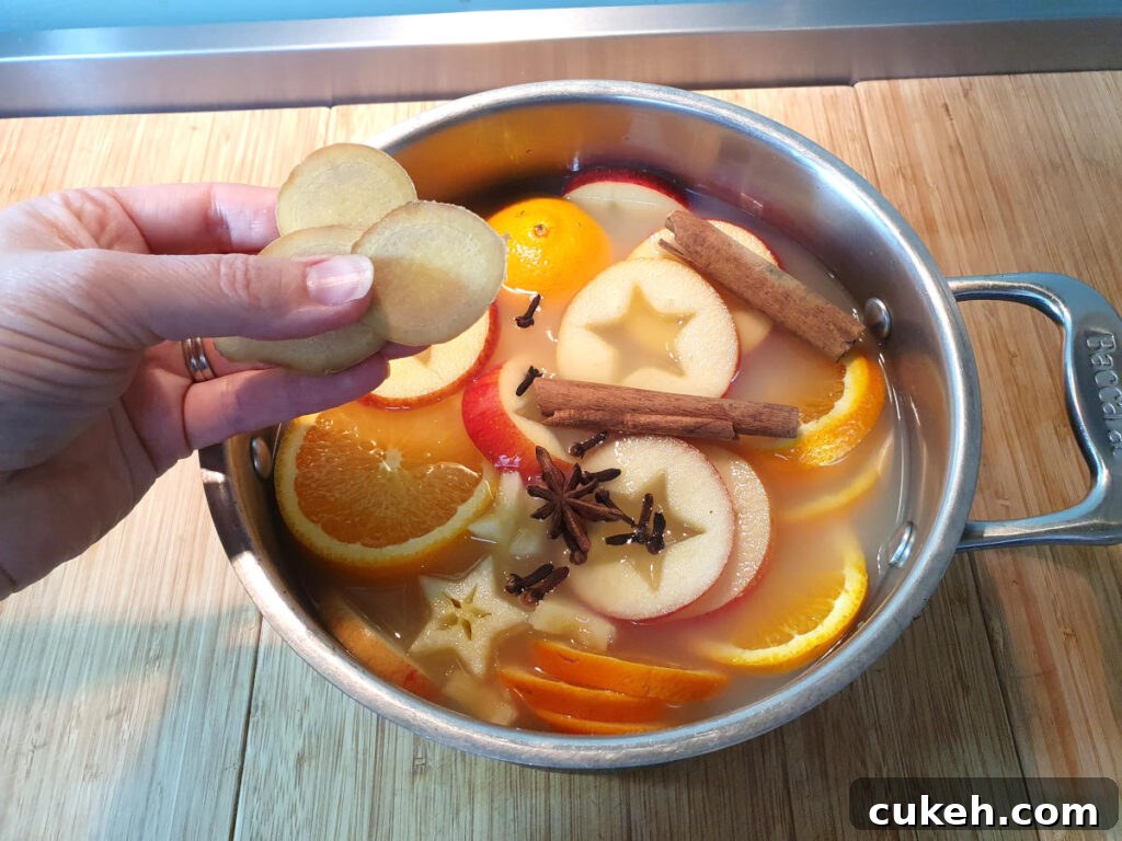 adding sliced ginger to a pot containing sliced apples and oranges, cloves, star annise and cinnamon sticks, on a chopping board.