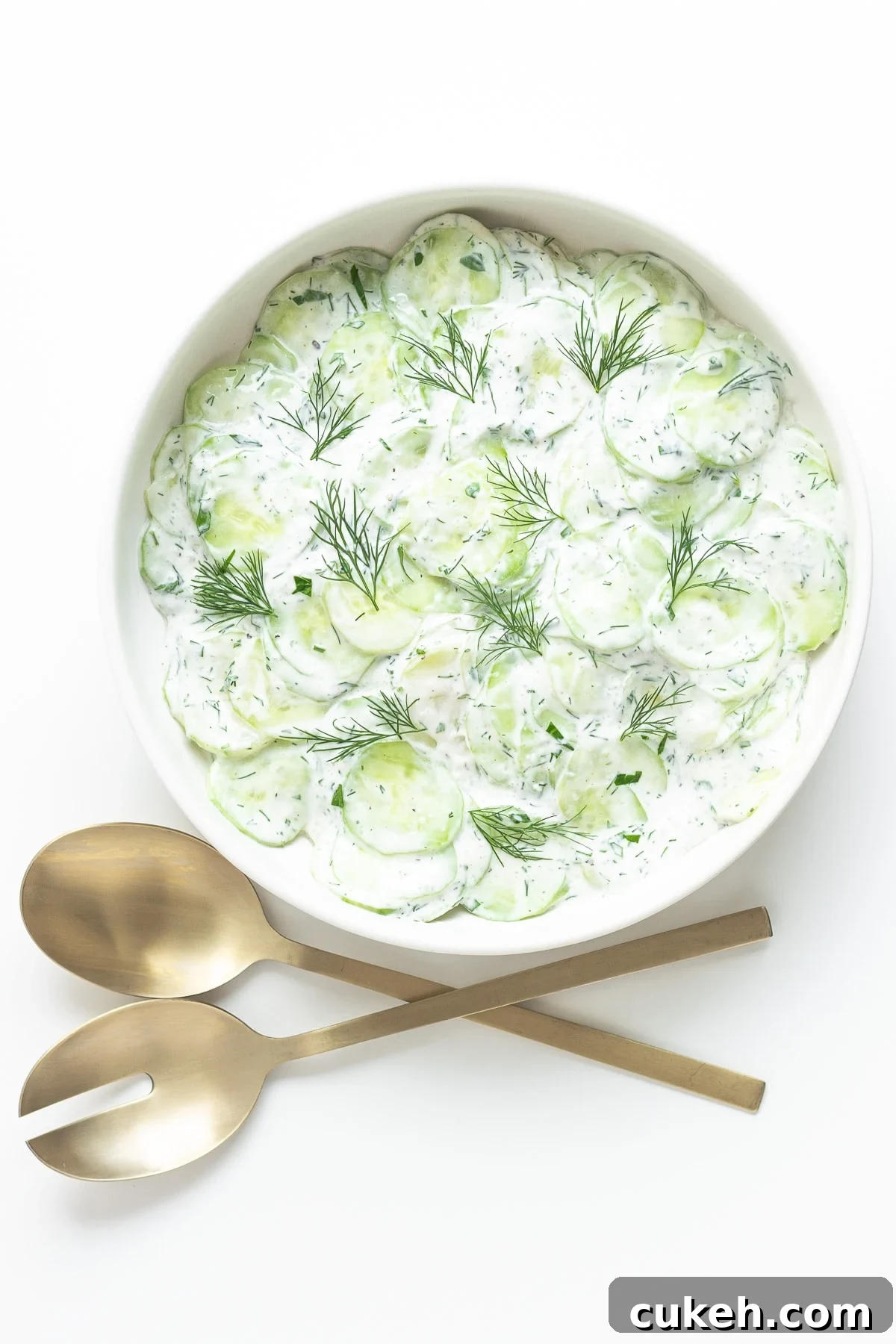 Cucumber salad in a white bowl with gold salad servers next to it.