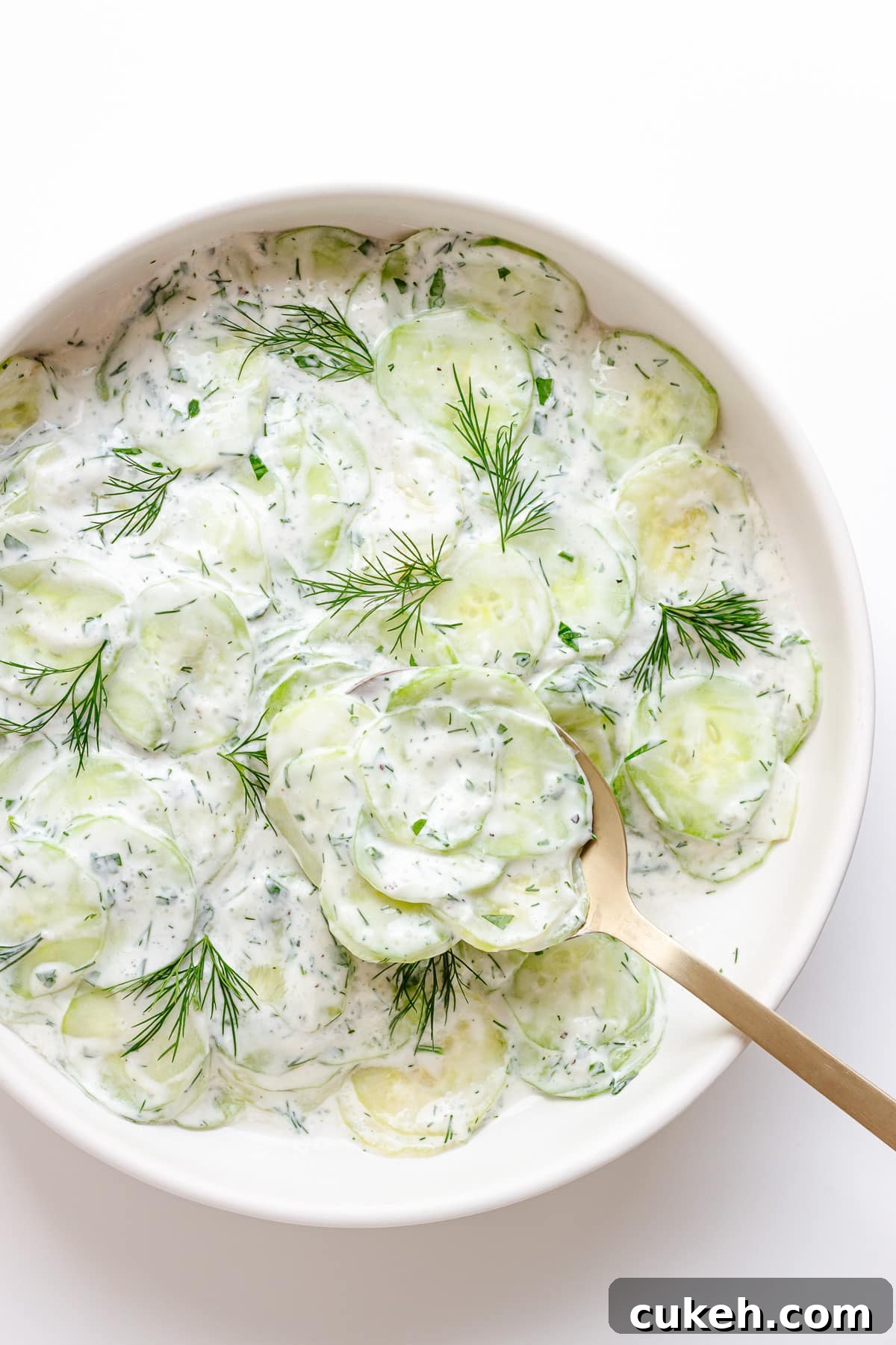 Gurkensalat (creamy German cucumber salad) in a white bowl with gold salad server lifting some out.