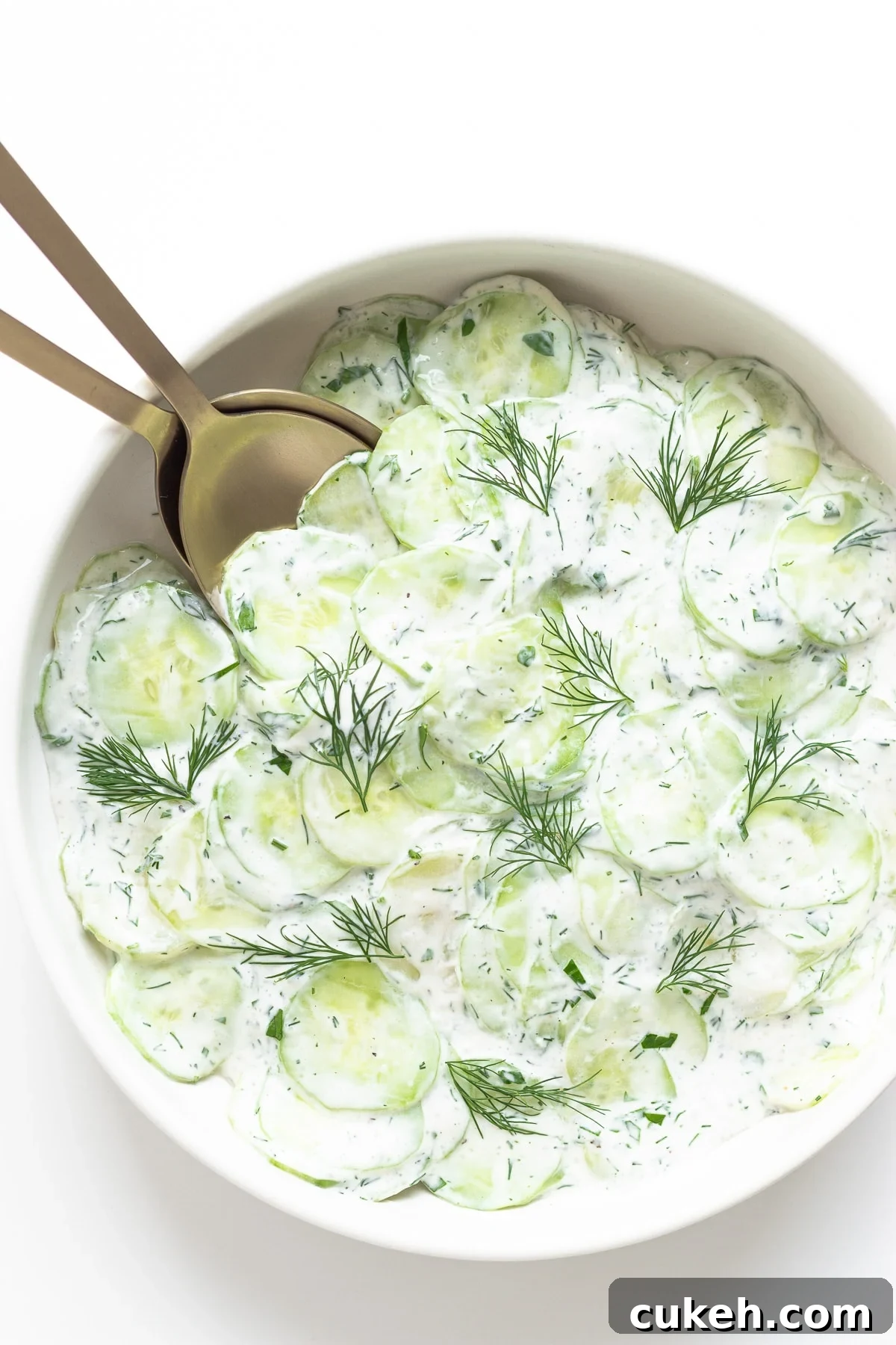 Sour cream cucumber salad in a white bowl with gold salad servers.