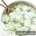 Close up of bowl containing creamy German cucumber salad (Gurkensalat) with gold salad servers.