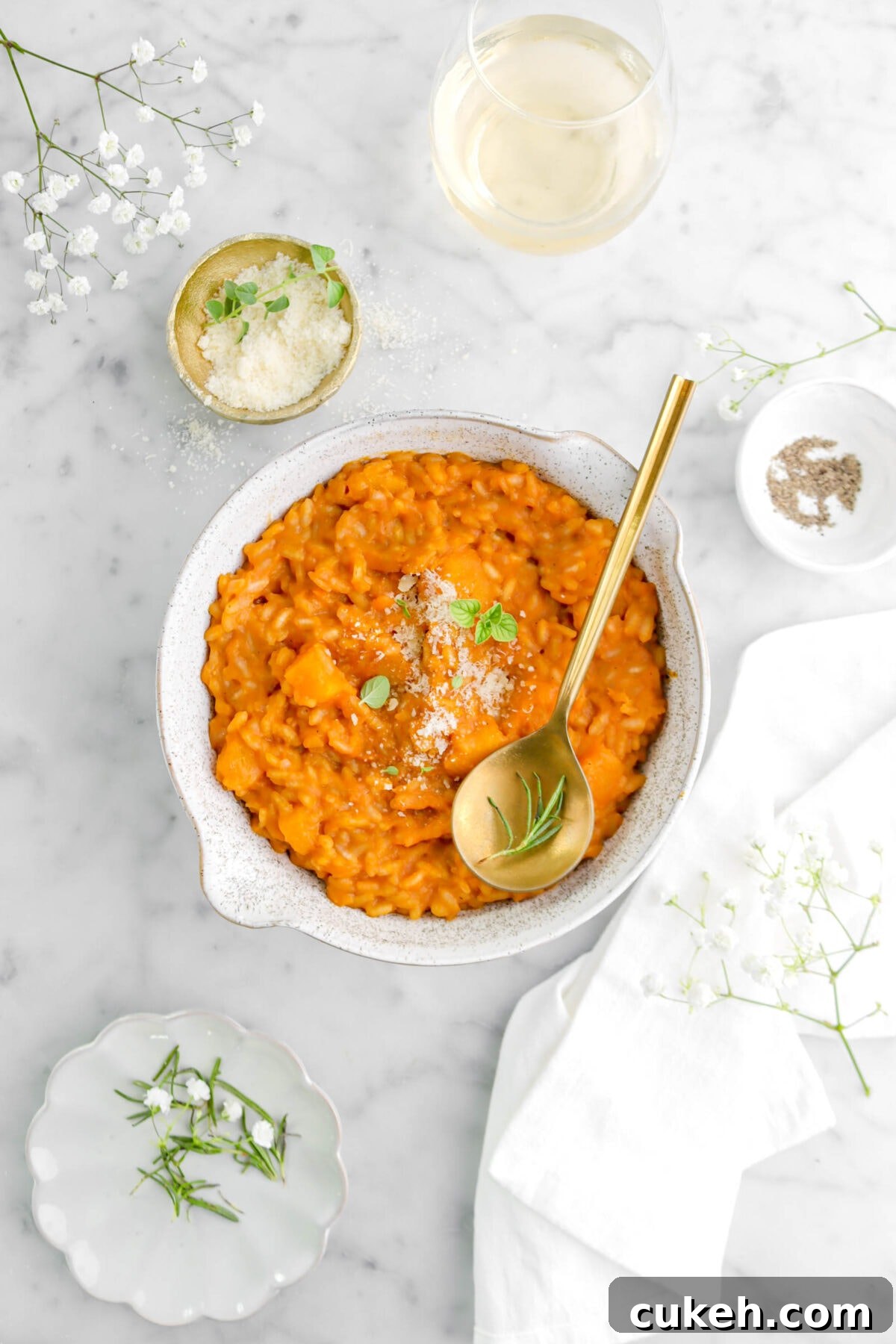 butternut squash risotto with cheese, oregano, gold spoon with rosemary sprig on top with white napkin and glass of wine beside.
