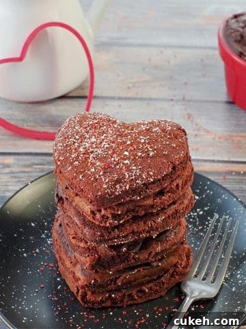 stack of heart-shaped red velvet pancakes on a black plate with a heart cookie cutter in the background
