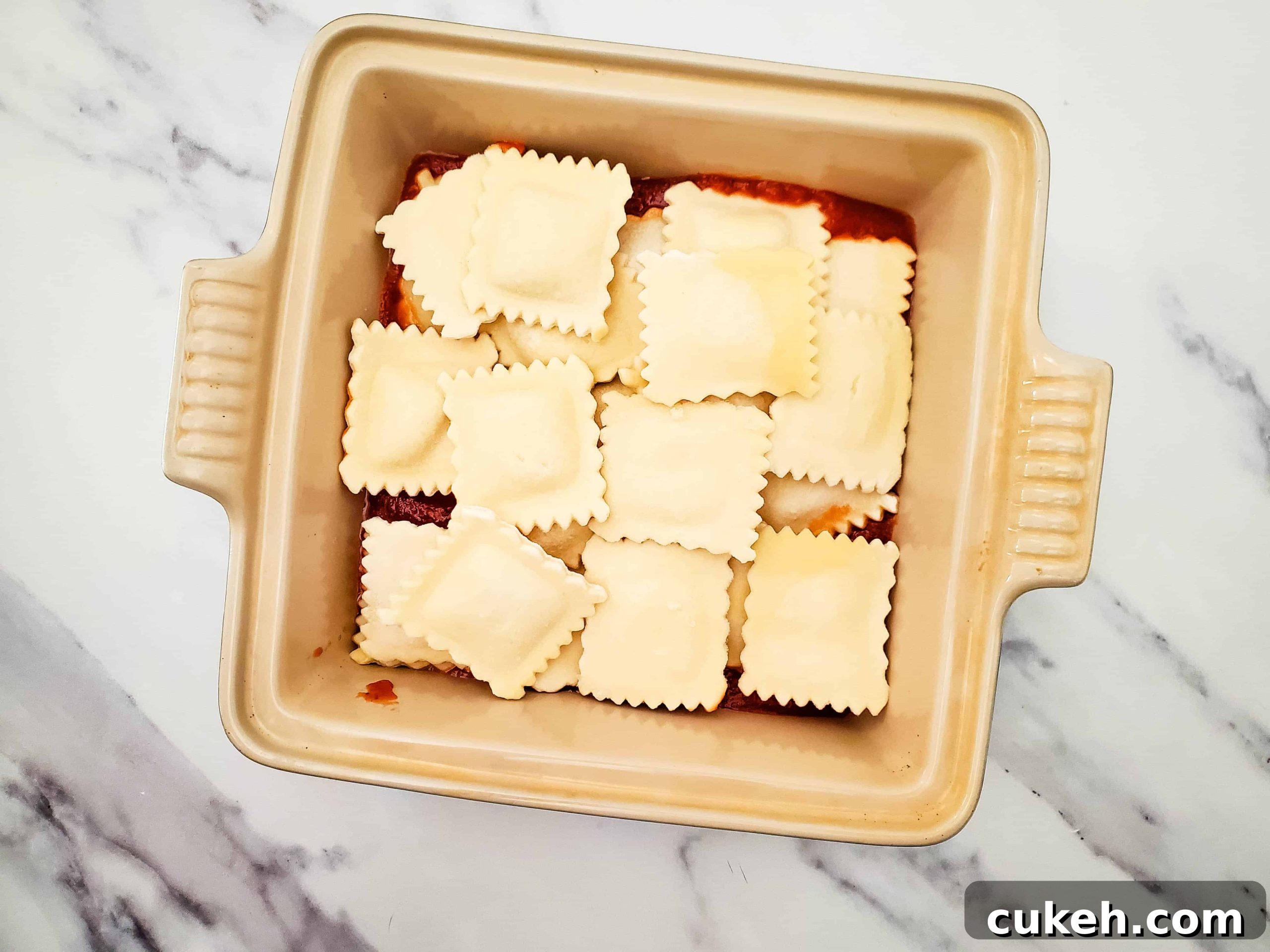 Glass baking dish with pasta sauce and frozen ravioli on top of a white counter