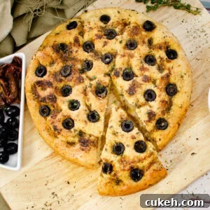 Olive Focaccia Bread on a wooden chopping board with slice cut out served with black olives and sun dried tomatoes on the side