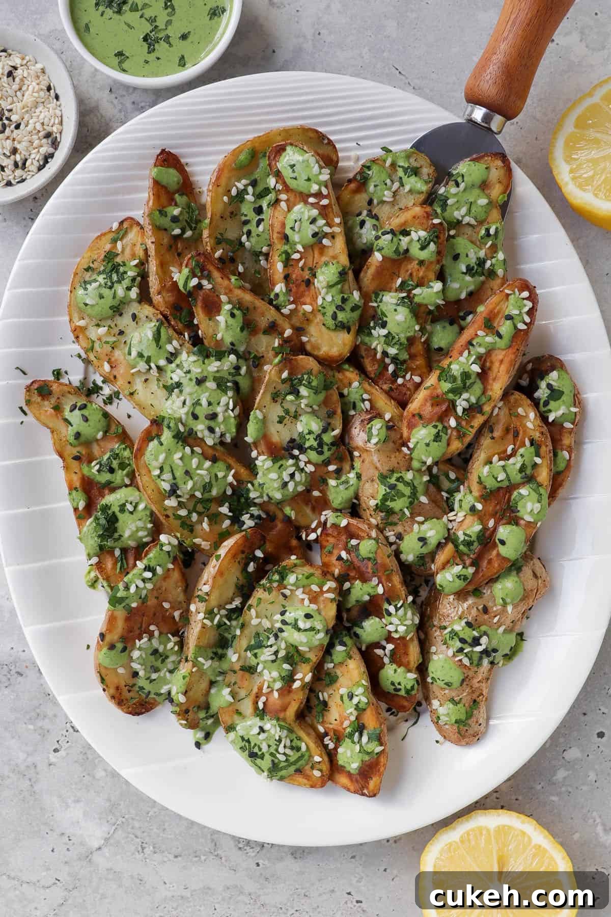 Air fryer fingerling potatoes on plate with green tahini sauce and serving spoon.. Lemons, extra tahini sauce, sesame seeds on the side.