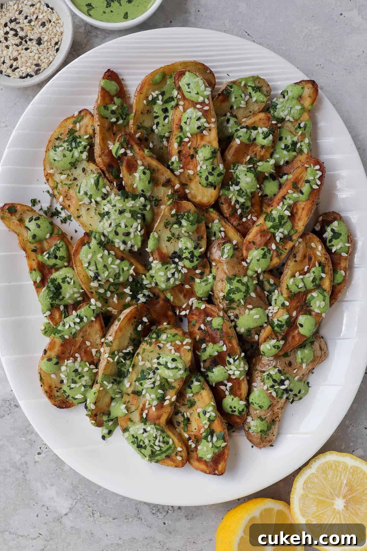 Air fryer fingerling potatoes on plate with herb tahini sauce drizzled on top. Lemons, extra tahini sauce, sesame seeds on the side.