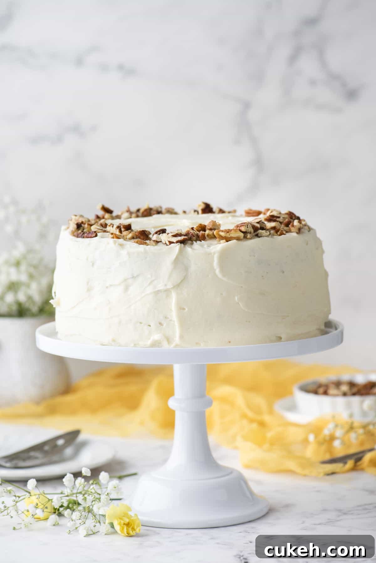 a hummingbird cake covered in cream cheese frosting and topped with chopped pecans on a white cake stand surrounded by white and yellow flowers, a yellow cloth and a bowl of pecans in the background