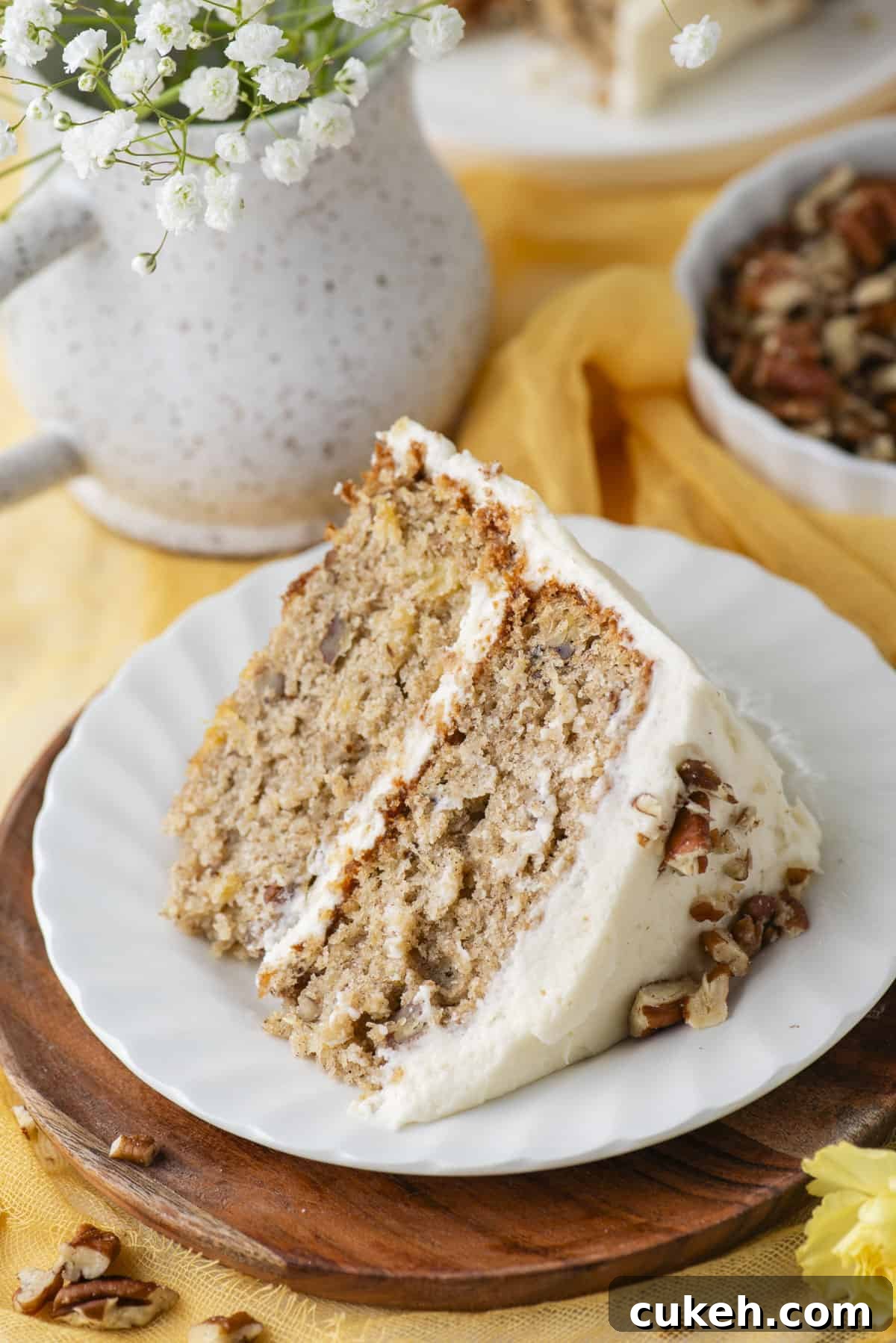 a slice of hummingbird cake on a small white plate on top of a round wood piece beside a dish of pecans and a vase of flowers
