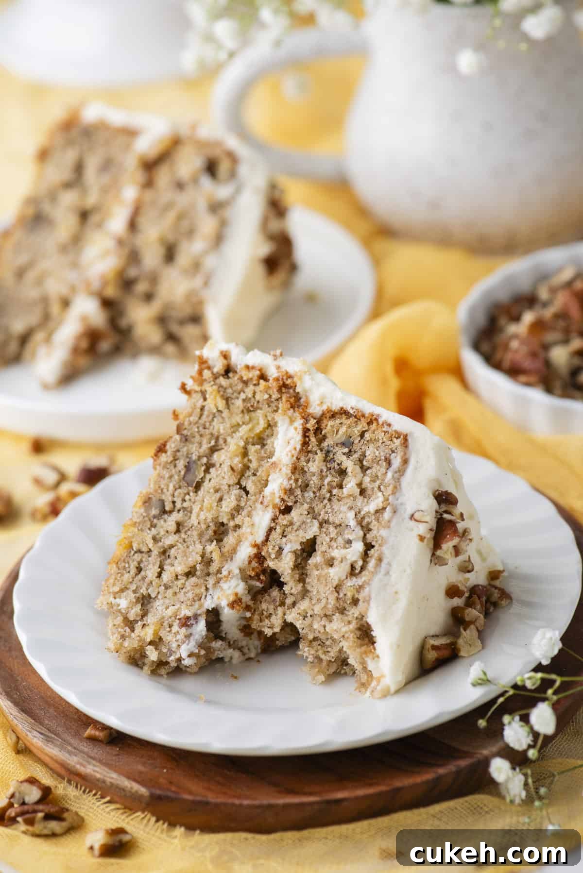 a slice of hummingbird cake on a white plate on top of a wooden circle, with one bite missing from the cake, chopped pecans sprinkled around, some white flowers in the corner and another plate with a slice of cake and a dish of pecans in the background