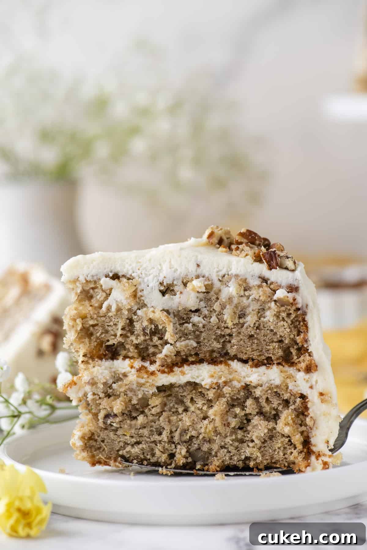 a slice of hummingbird cake being placed on a white plate with a spatula, showing the side of the two layers of cake with cream cheese frosting in the middle, with white and yellow flowers on the left