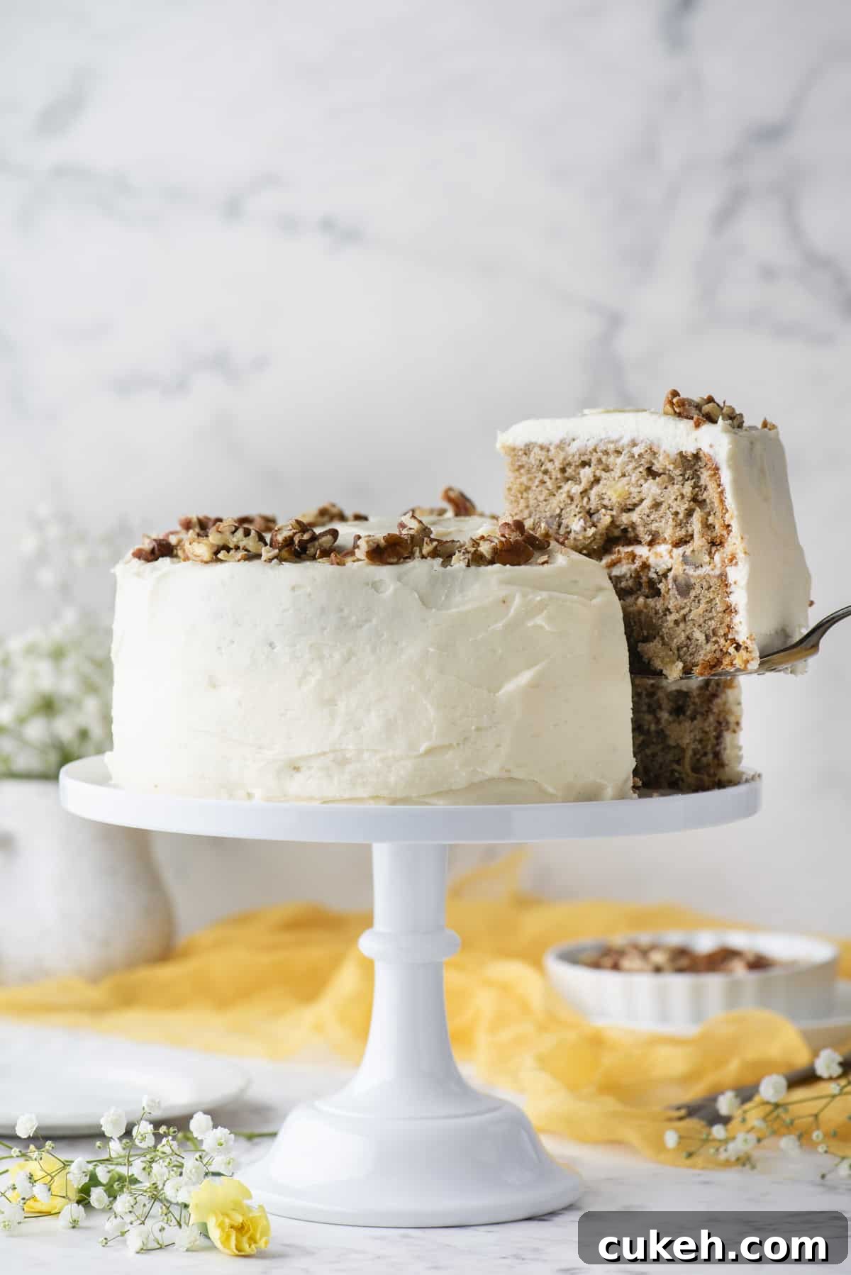a hummingbird cake on a white cake stand with a piece of the cake being lifted away with a spatula, beneath it white and yellow flowers, a yellow cloth and a white dish of pecans