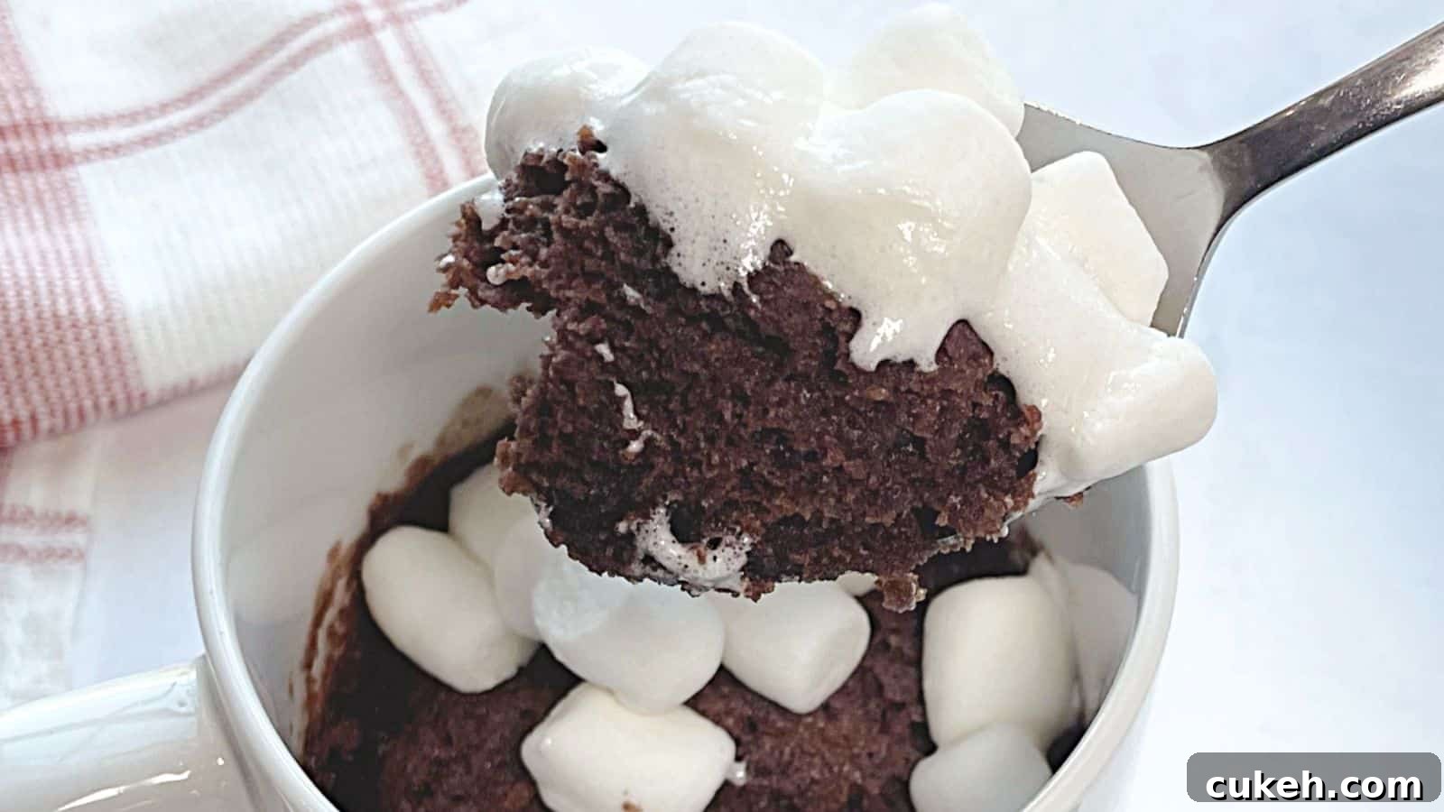 A spoon lifting a bite of chocolate mug cake topped with melted marshmallows from a white mug.