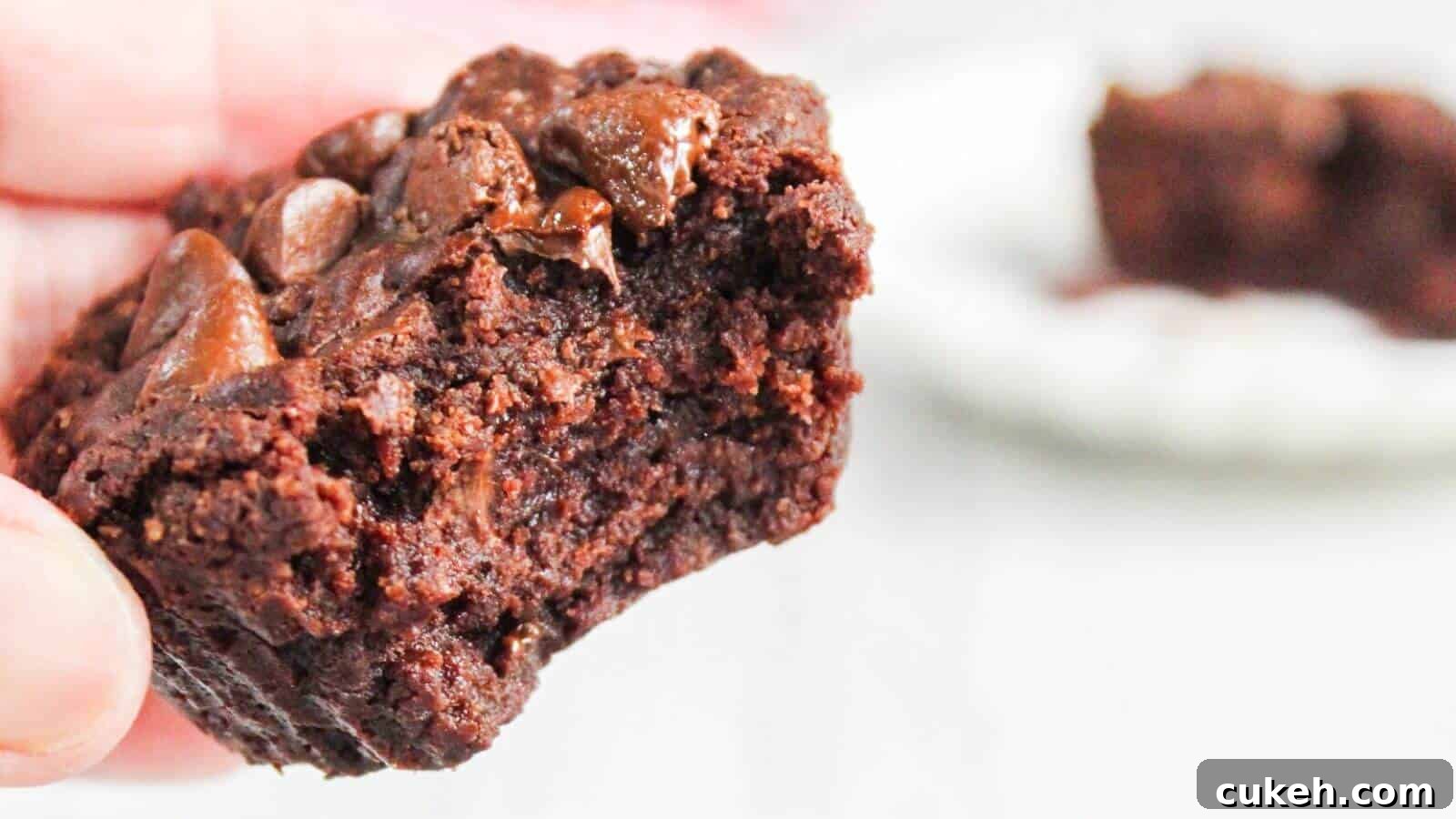 Close-up of a hand holding a half-eaten chocolate brownie with gooey chocolate chunks, with another partial brownie blurred in the background.