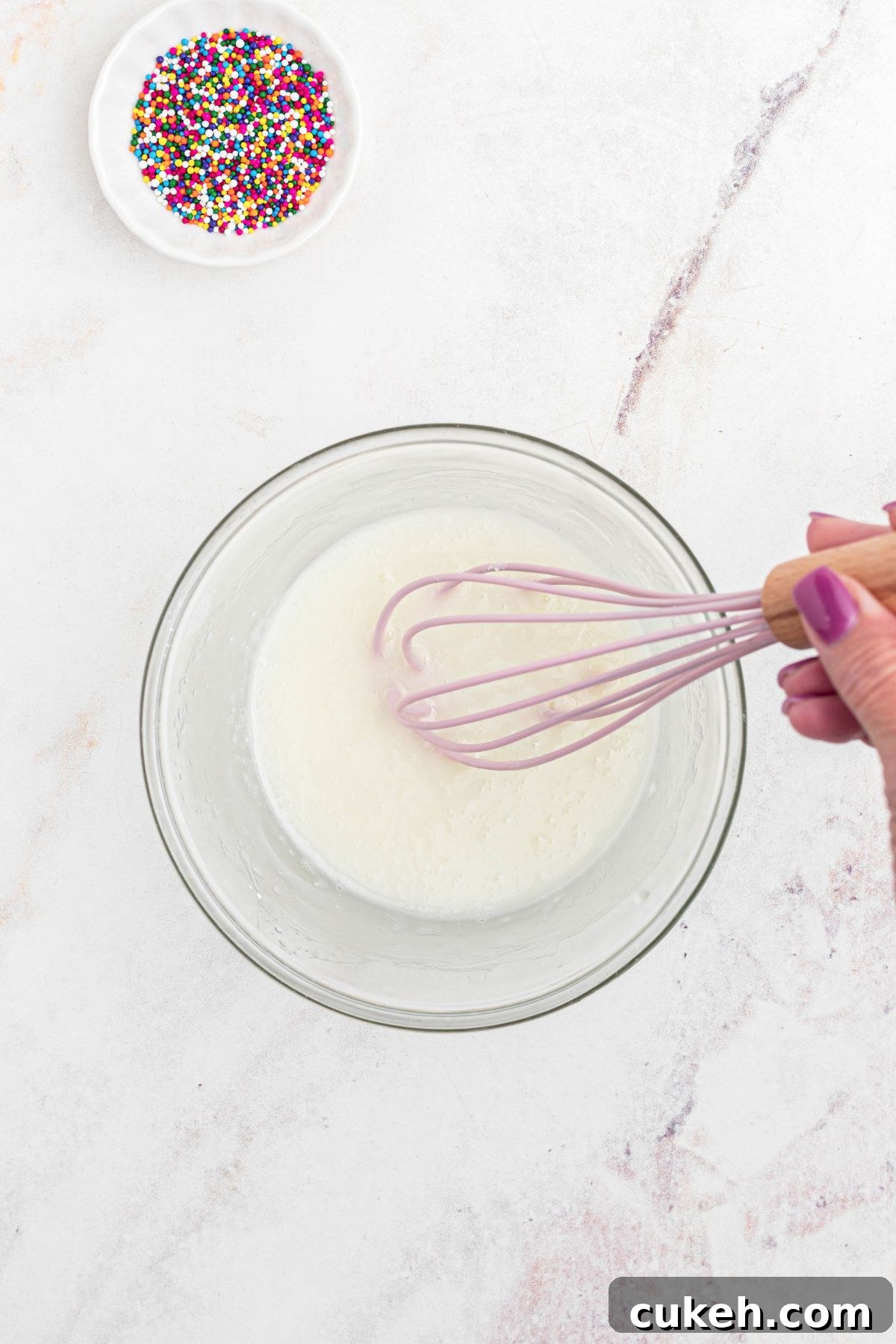 Clear glass bowl with powdered sugar and milk being mixed together, on a marble table. 
