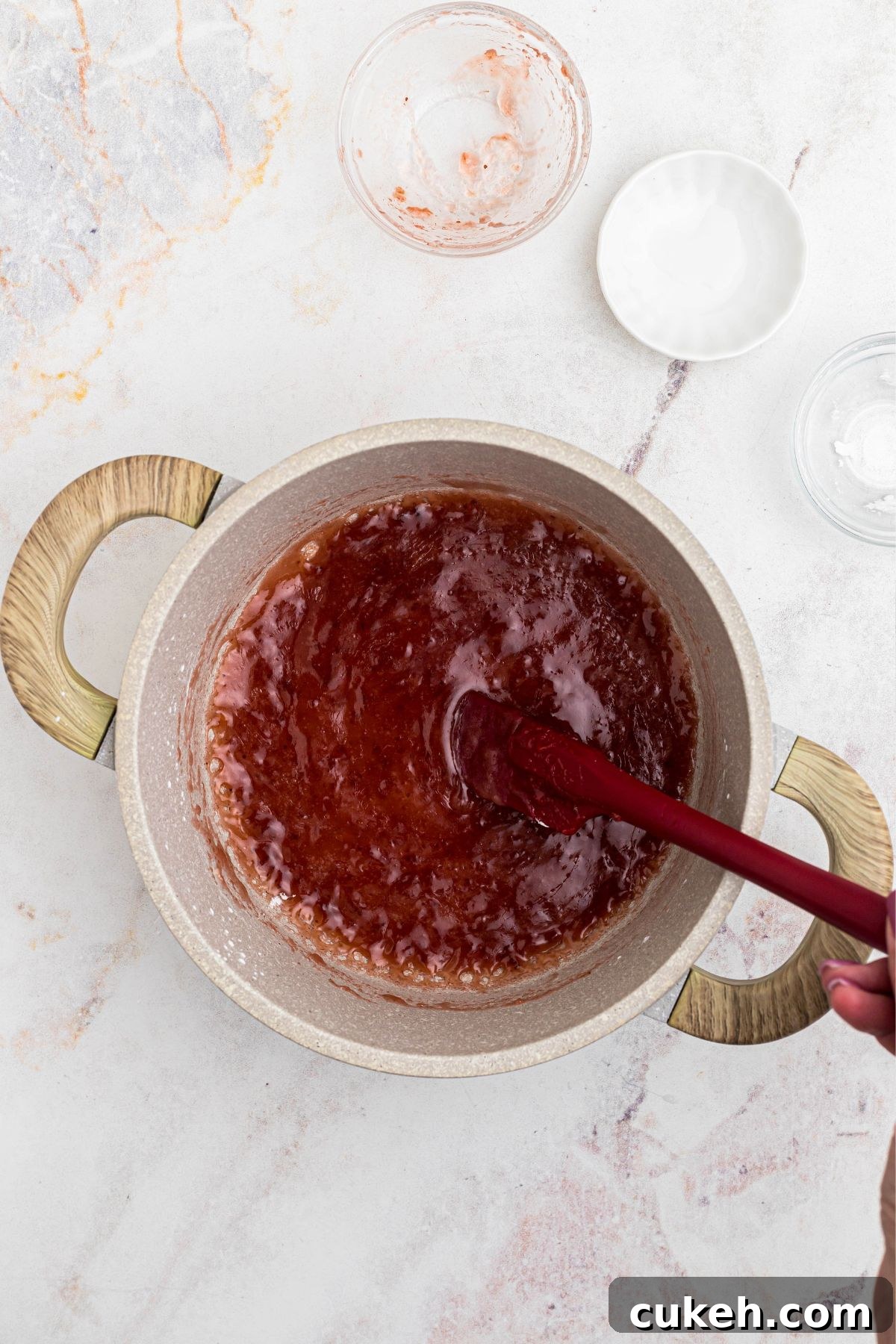 Jam thickened in a saucepan, being stirred by a red spatula. 