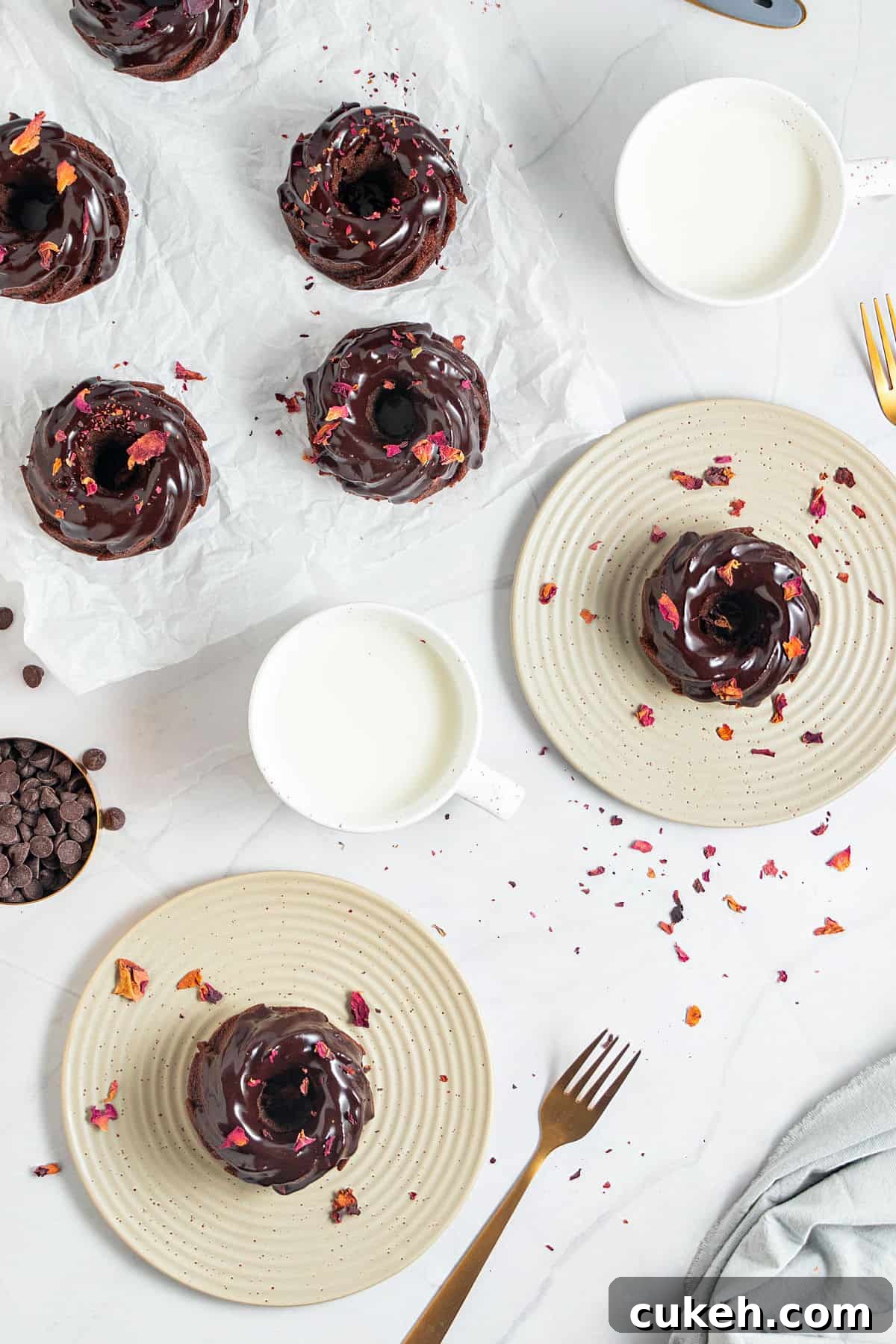 Mini Chocolate Bundt Cakes with Chocolate Glaze 3 Top view of white surface with two plates and white paper with glazed chocolate mini bundts. Rose petals, forks, milk glasses.