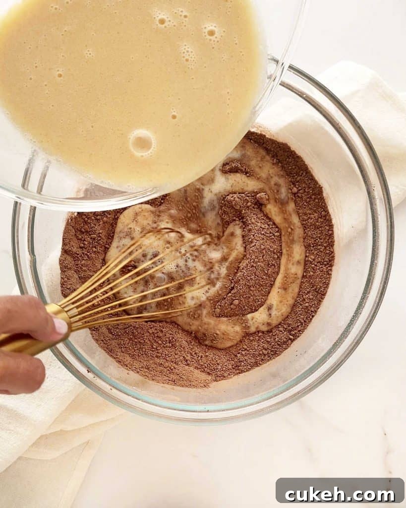 Mini Chocolate Bundt Cakes with Chocolate Glaze 5 Adding wet ingredients into chocolate cake dry ingredients in a glass bowl on a white surface.