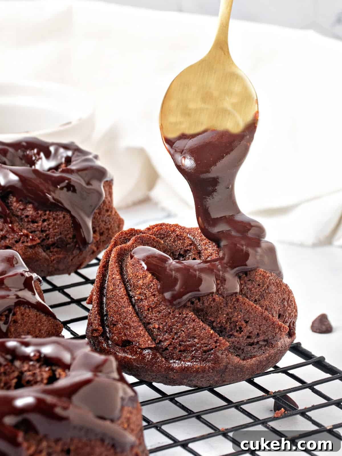 Mini Chocolate Bundt Cakes with Chocolate Glaze 9 Glazing chocolate mini bundt cakes with ganache. Golden spoon, wire rack. White background.