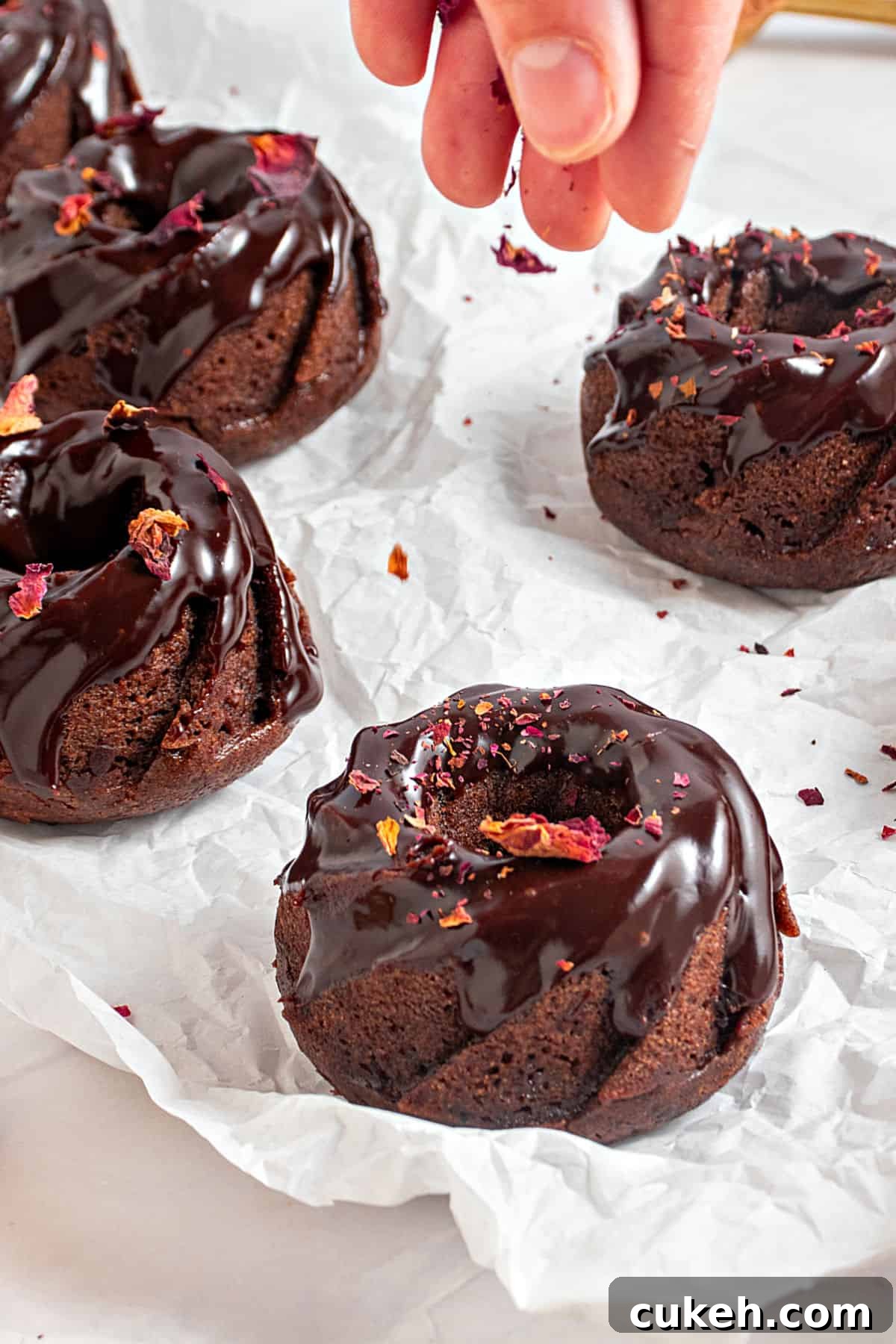 Mini Chocolate Bundt Cakes with Chocolate Glaze 10 Sprinkling rose petals on chocolate glazed mini bundt cakes. White paper surface.