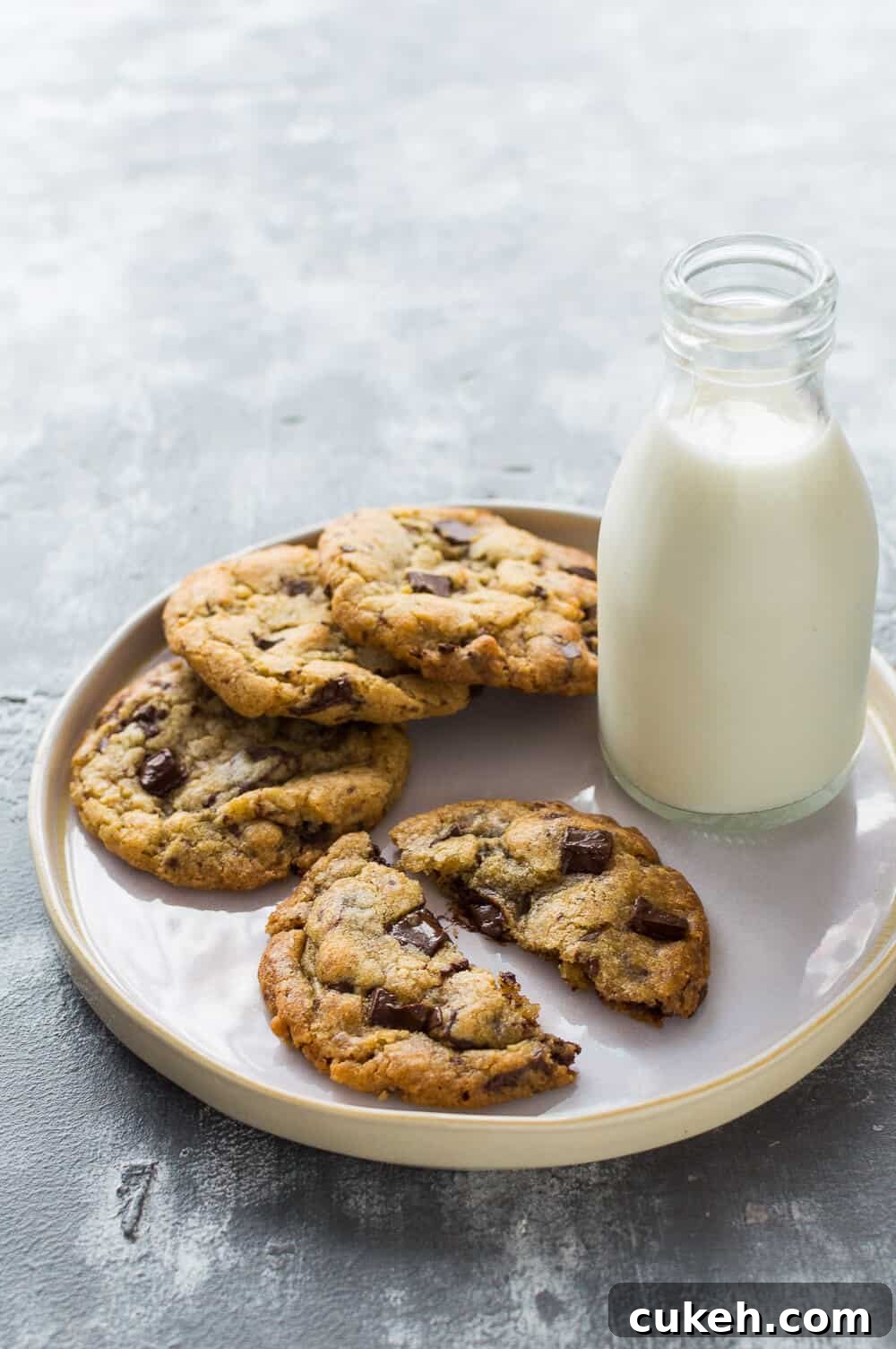 Photo of a white plate of vegan chocolate chip cookies with a glass bottle of almond milk on a grey background
