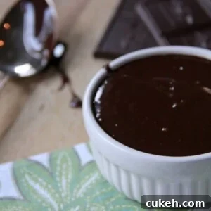 A white ramekin filled with rich chocolate ganache sits on a patterned napkin, with a spoon and pieces of chocolate in the background.