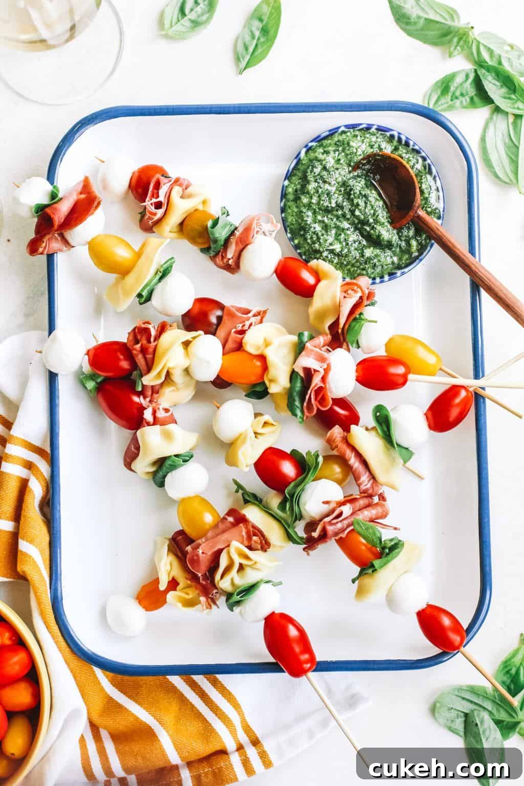 Tomato, mozzarella, basil, Prosciutto, and tortellini skewers with a bowl of pesto in white casserole dish