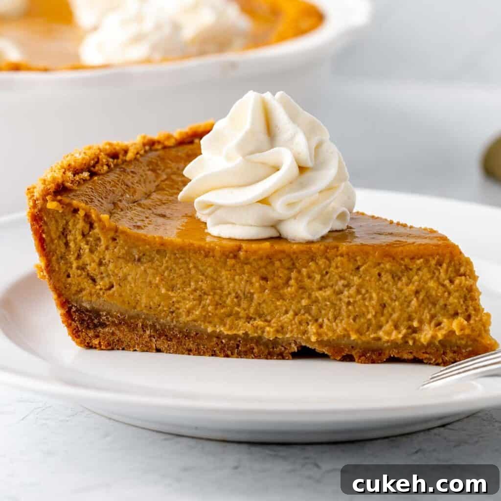 A slice of pumpkin pie on a white plate, topped with whipped cream. A white pie plate and the remaining pie in the background.