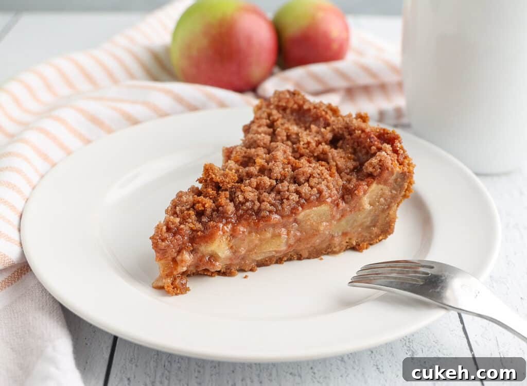 A slice of apple pie on a white plate with a fork to the side and apples in the background.
