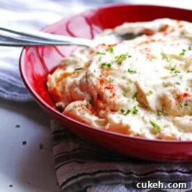 Baked mashed potatoes in a red bowl with a spoon. The potatoes are garnished with chives.