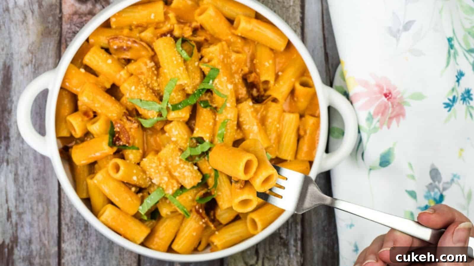 A white bowl of rigatoni pasta with creamy sauce, garnished with cheese and green herbs, is placed on a wooden surface. A hand is holding a fork with a piece of pasta.