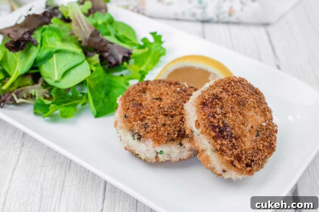 Two Salmon Cakes on a white plate with mixed leafy greens and a lemon wedge.