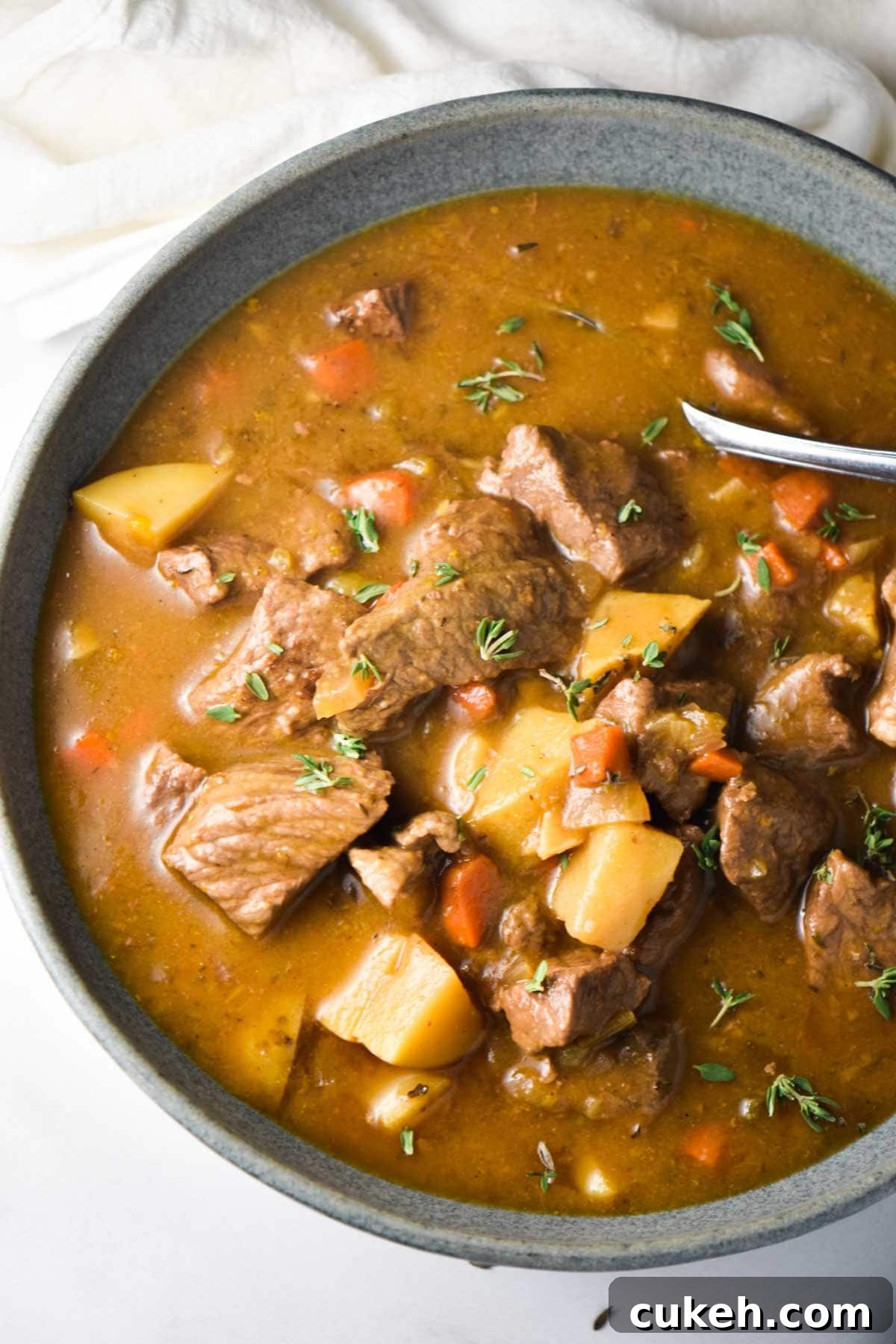 A close up of beef chuck roast, potatoes, carrots, and thyme in a stew with a spoon dipping in